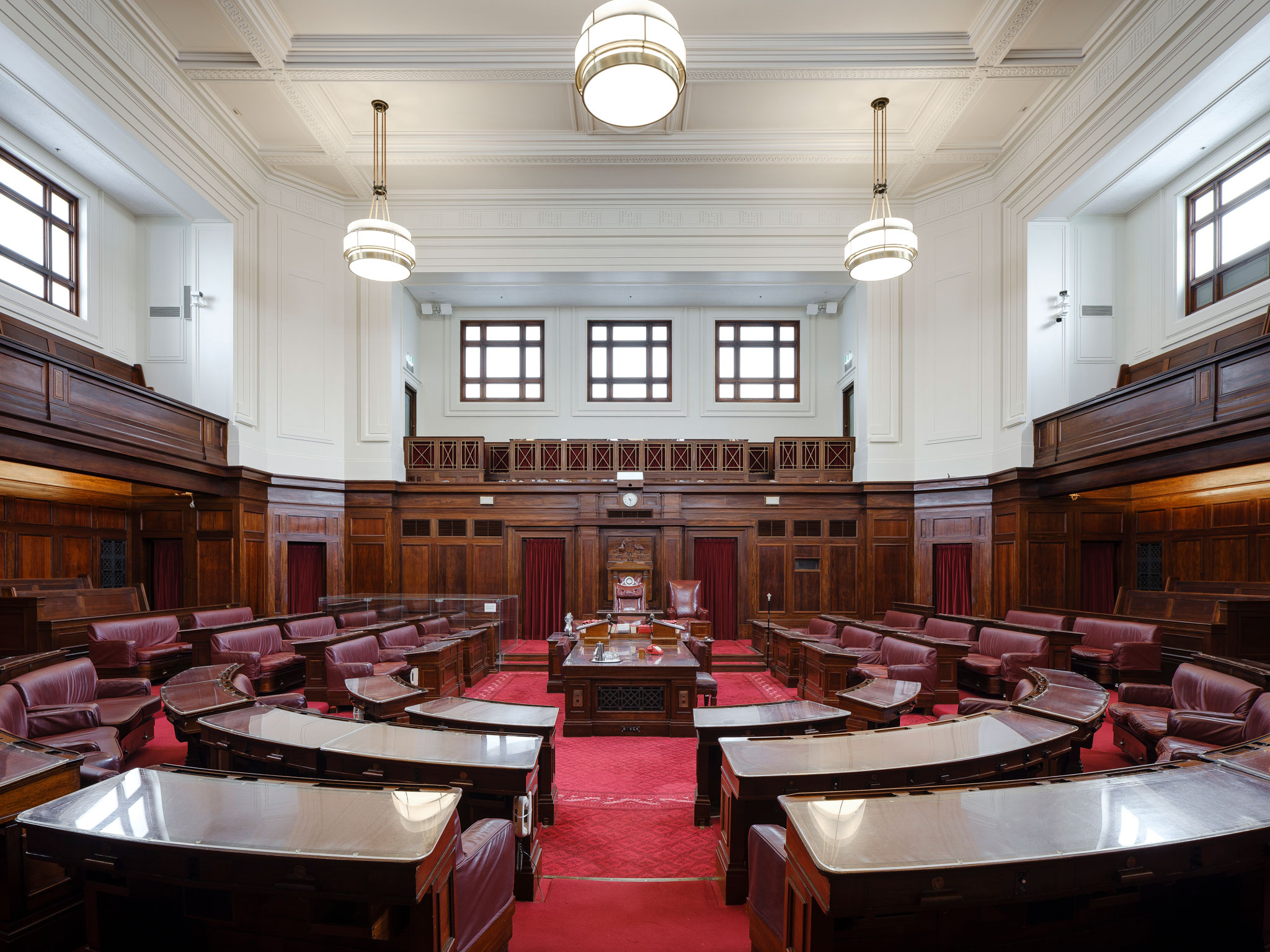 A view into the Senate Chamber looking straight into the room with benches in a U-shape on either side of the centre table. The carpet is a rich red and the seats are red leather. Art deco lights hang from the ceiling and wood timber panels surround the room. 