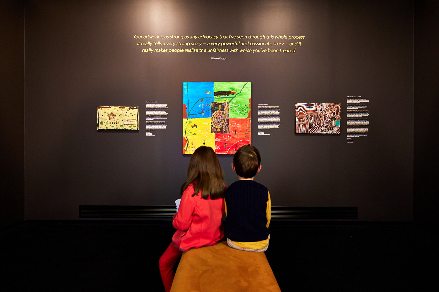 A boy and girl sit on a stool looking up at artworks from Jack Green. 