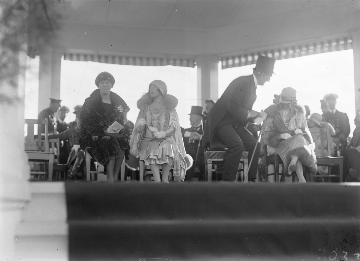 Mrs Ethel Bruce and the Duchess of York chat as Prime Minister Stanley Melbourne Bruce takes his seat on the Royal dais next to Lady Stonehaven, the wife of the Governor-General.
Credit: Photograph by William J Mildenhall NAA: A3560, 3033 This black and white photograph lets us peek over the edge of the carpeted floor of the white timber Royal dais constructed at York Park for the official party to watch the Troop Review. On the left, a seated Mrs Ethel Bruce chats with the Duchess of York. Ethel is dressed in a dark, luxurious fur coat with cloche hat and gloves. The Duchess is richly dressed in a pale-coloured lustrous dress with elaborate embroidery and a fur-trimmed cape, cloche hat and white gloves. Prime Minister Bruce, in top hat and
