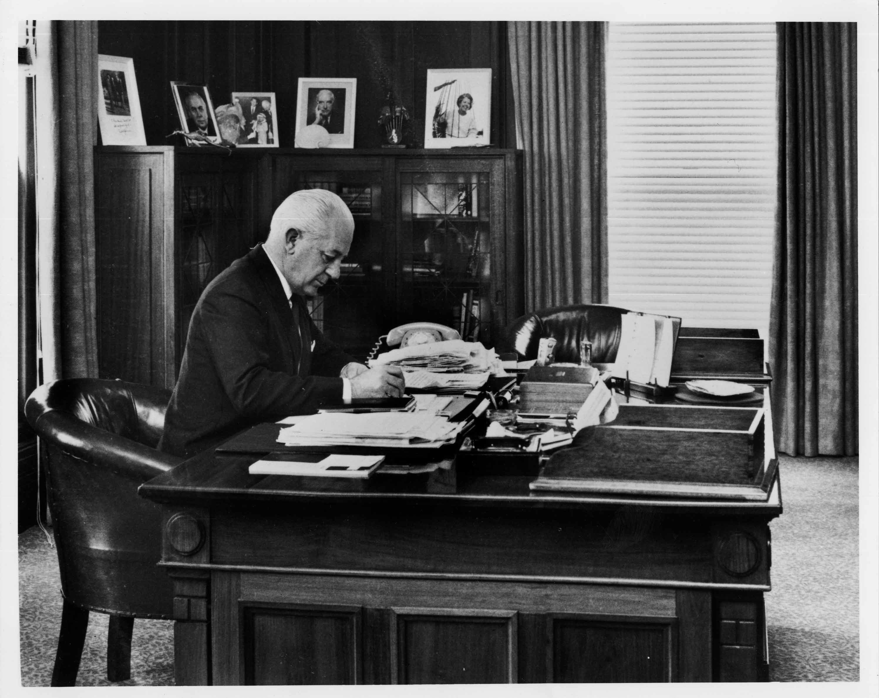 In this black and white photograph Prime Minister Harrold Holt sits at the timber desk made in 1926. Holt is wearing a dark suit, white shirt and tie and is leaning on the desk while he writes. The desk has fine carved panels with a geometric detailing on the corners. The surface of the desk is cluttered with stacks of documents and a telephone. On the glass-fronted cabinets in the background are a number of framed photographic portraits.  