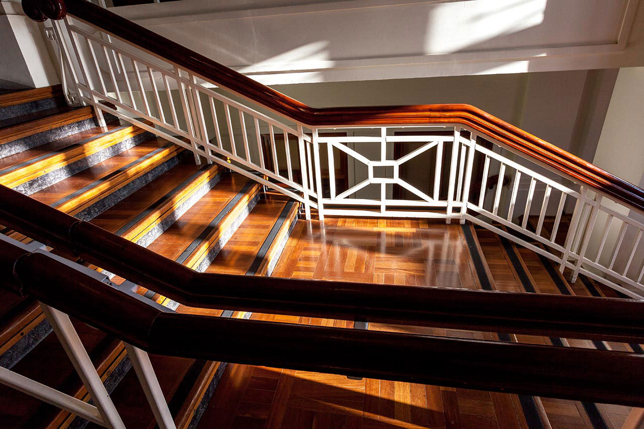 A close up of a timber staircase with dappled light. The railings feature white posts with an art-deco style design.