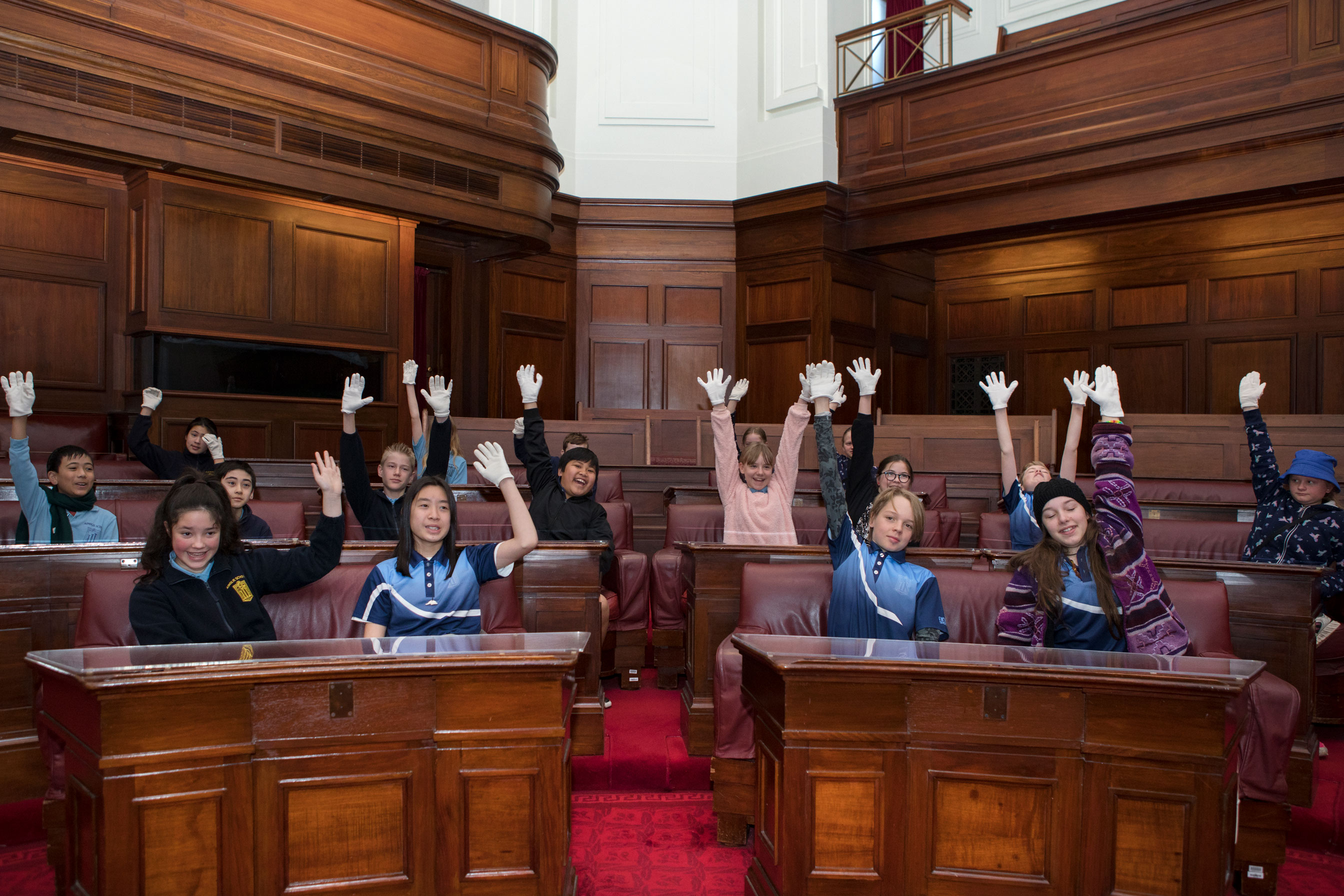 School children raise their hands while sitting in the Senate Chamber which has red carpet and wood panelled benches in a U-shape. 