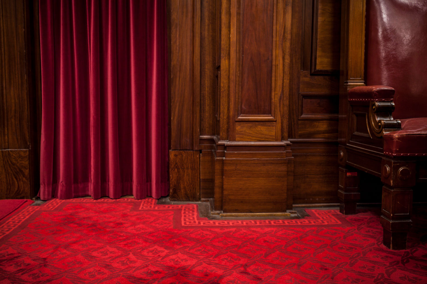 Close up of a red velvet curtin falling to a red carpeted floor with a timber furniture panel next to it.