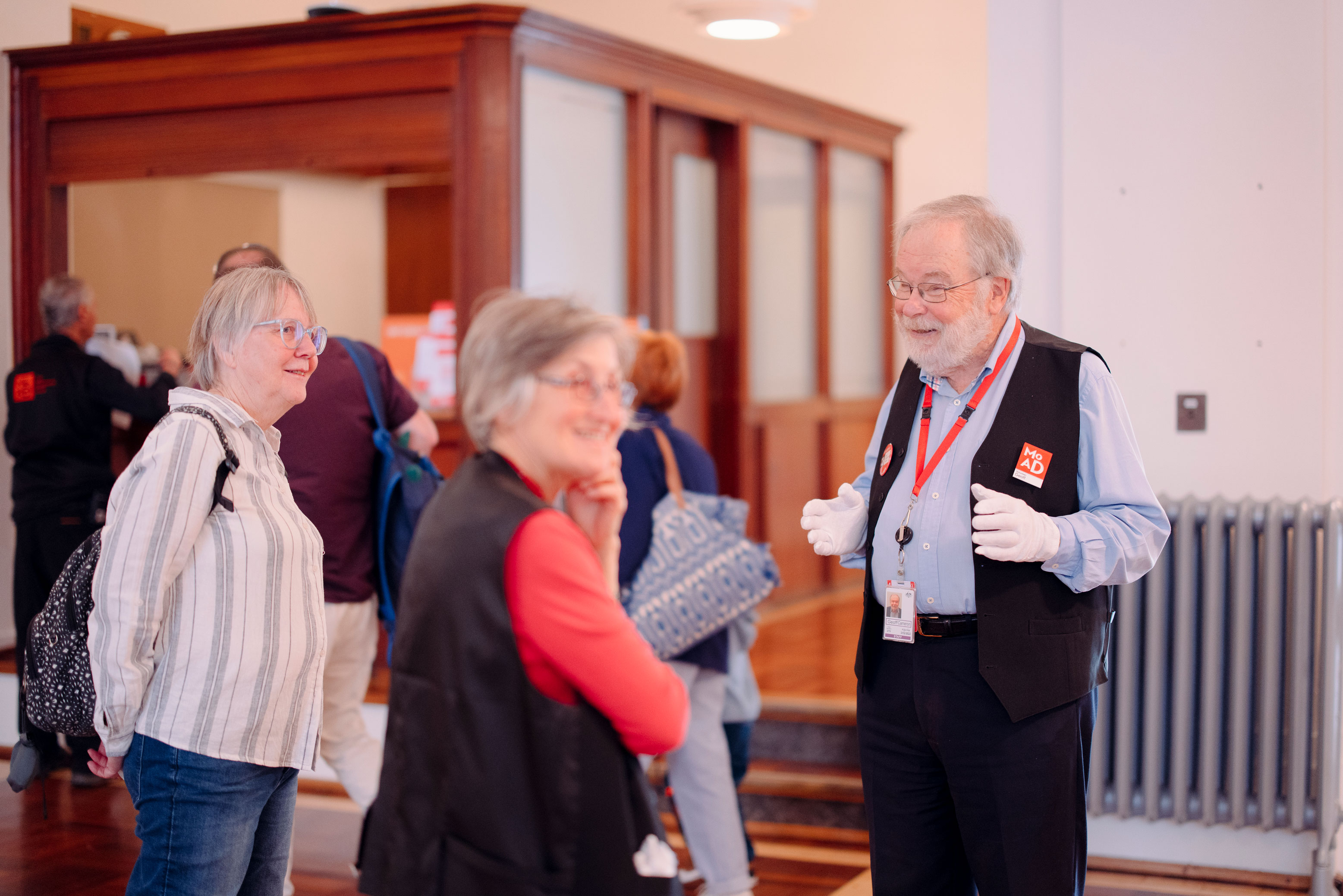 A MoAD volunteer smiles wearing white gloves and talking to two visitors. 