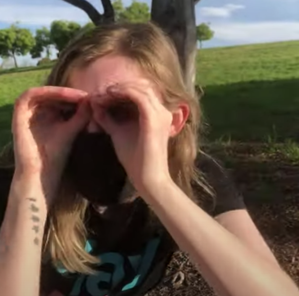 A woman stands outside with her hands up to eyes like binoculars. 