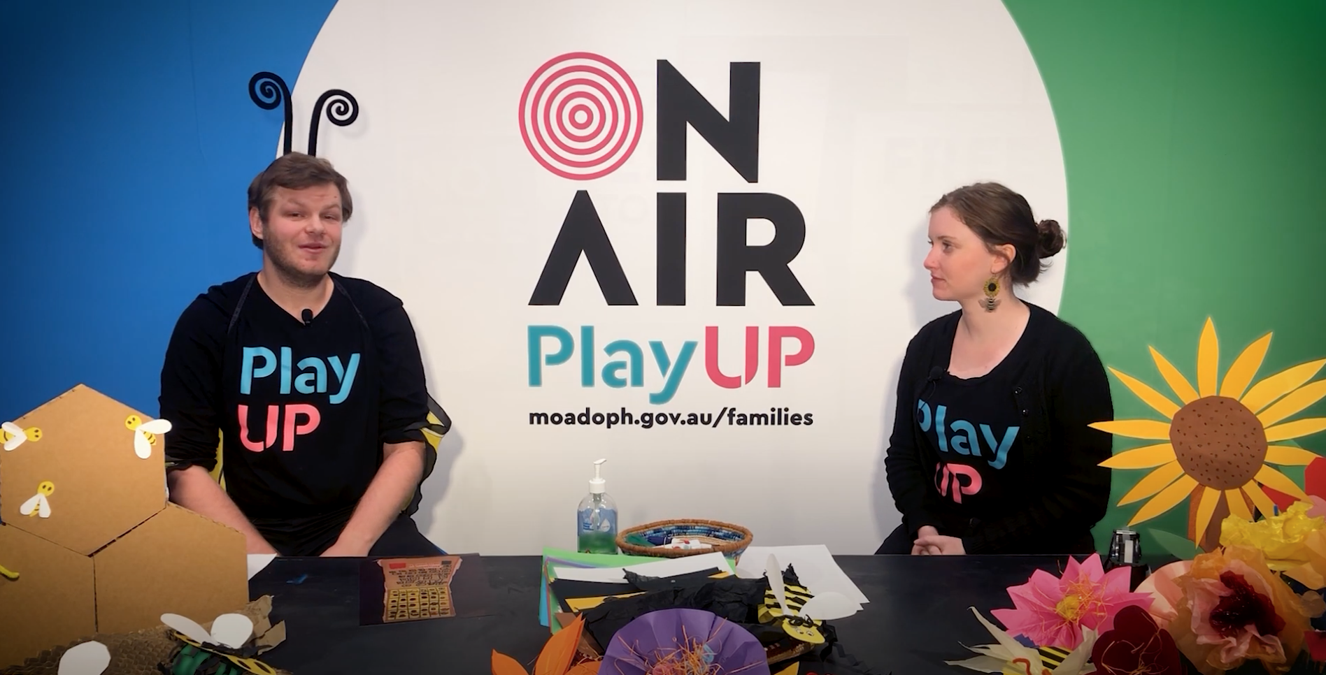 Two PlayUP facilitators stand behind a table covered in craft supplies. One wears a headband with bee feelers.