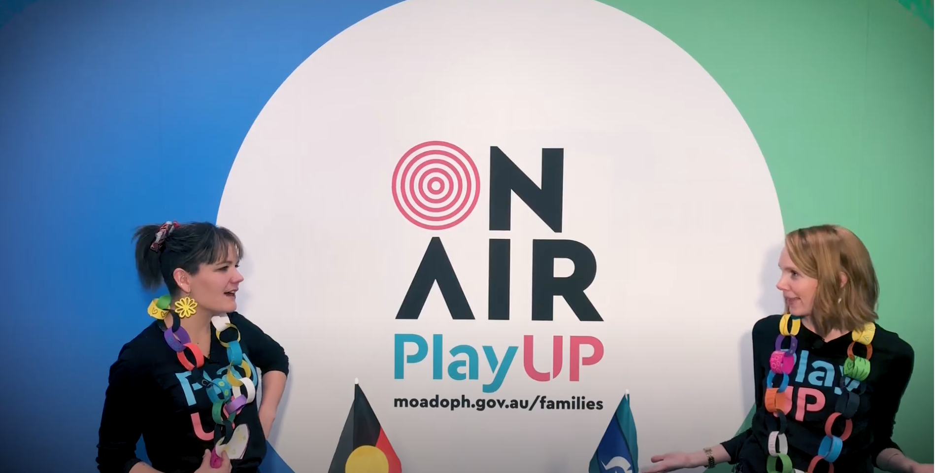 Two PlayUP facilitators wearing black shirts that say 'PlayUP' stand behind a table covered in craft supplies.