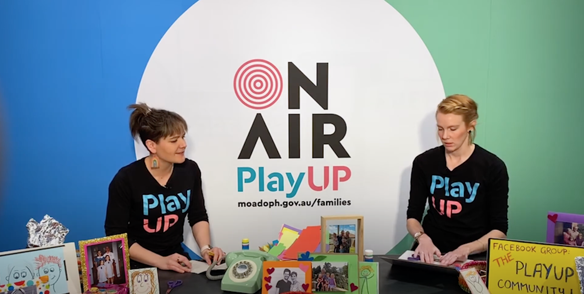 Two PlayUP facilitators stand behind a table covered in craft supplies. 