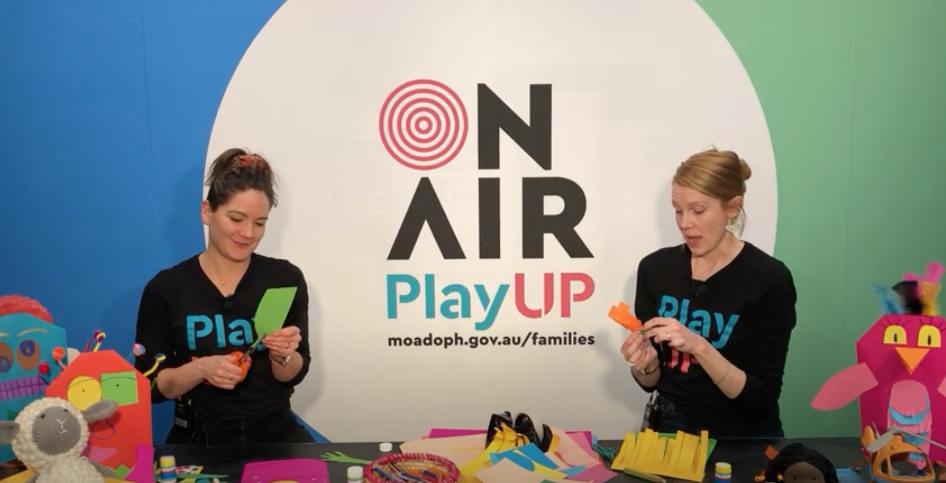 Two PlayUP facilitators cut cardboard behind a table filed with craft supplies. 