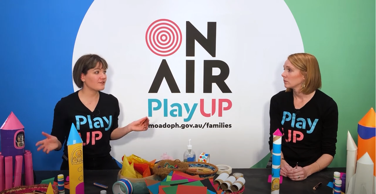 Two facilitators wearing black shirts that say PlayUP make something out of cardboard.