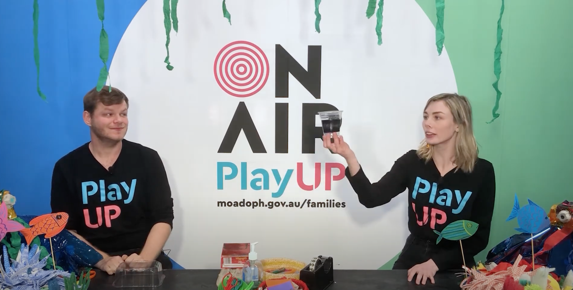 Two PlayUP facilitators stand behind a table covered in craft supplies. 