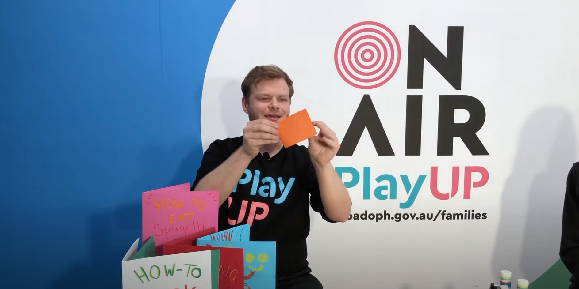 A PlayUP facilitator holds a piece of cardboard. There are handmade books made out of cardboard on the table in front of him.