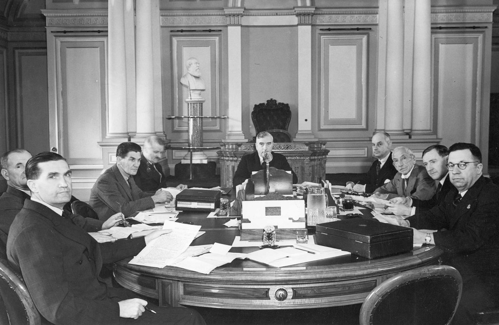 Nine men are gathered, seated around a large table covered with paper documents, with Robert Menzies seated at the head of the table.