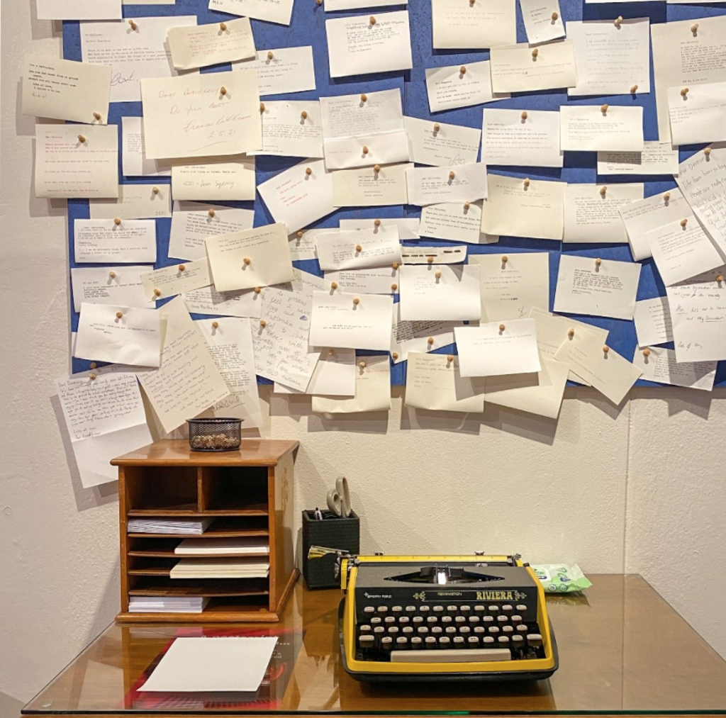 A yellow typewriter sits on a desk in front of a pinboard covered in typewritten notes.