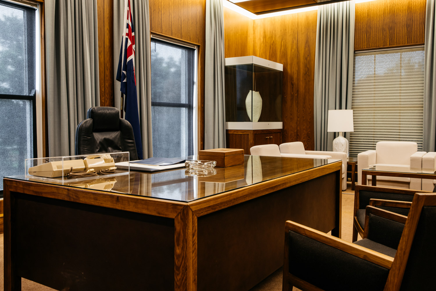 A colour photograph of the Prime Minister's Desk. Behind it is a comfortable office chair and an Australian flag on a pole. In front and to the side of the desk are armchairs.