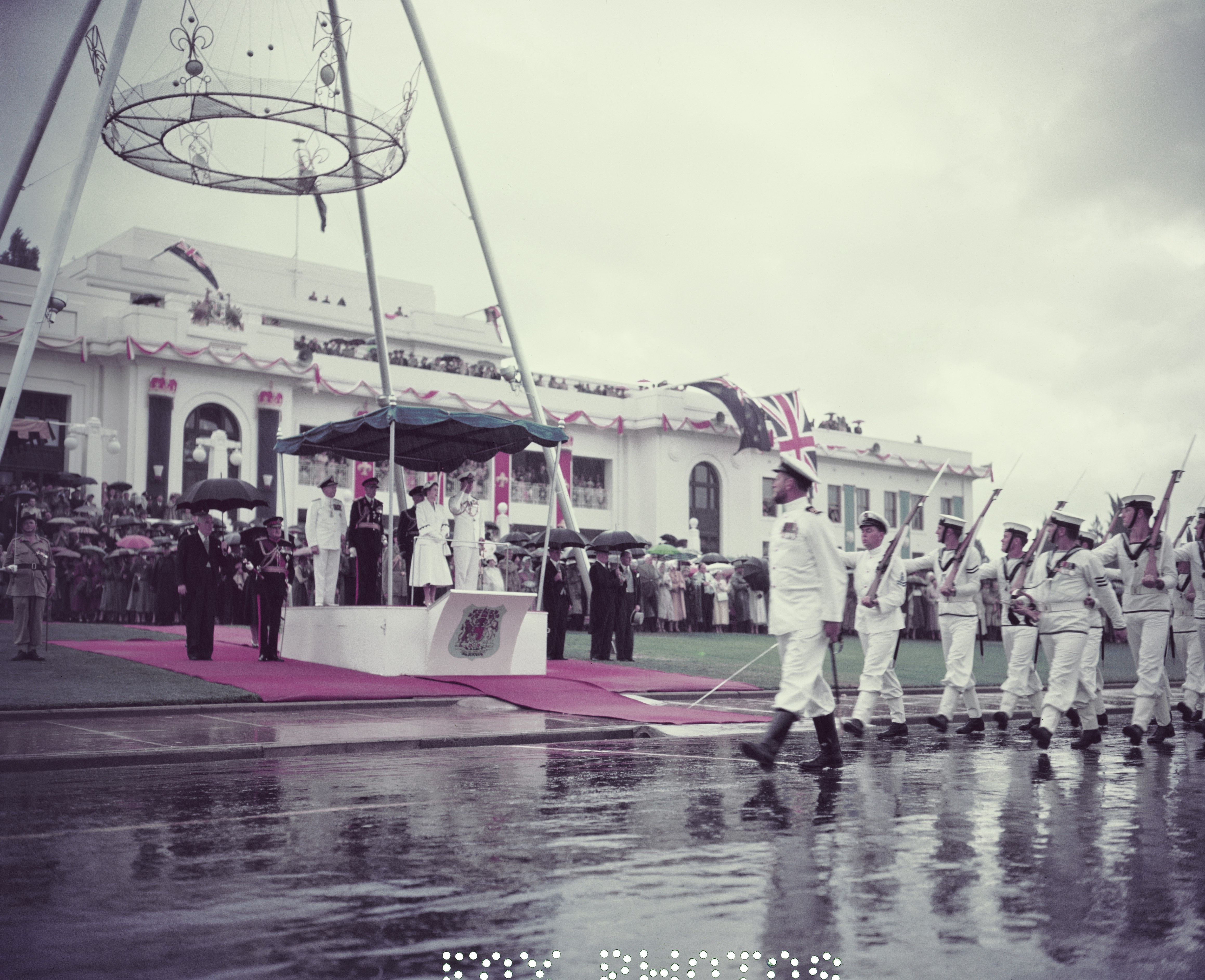 Queen Elizabeth II stands on a podium in front of a crowded (Old) Parliament House, as a naval procession marches past.