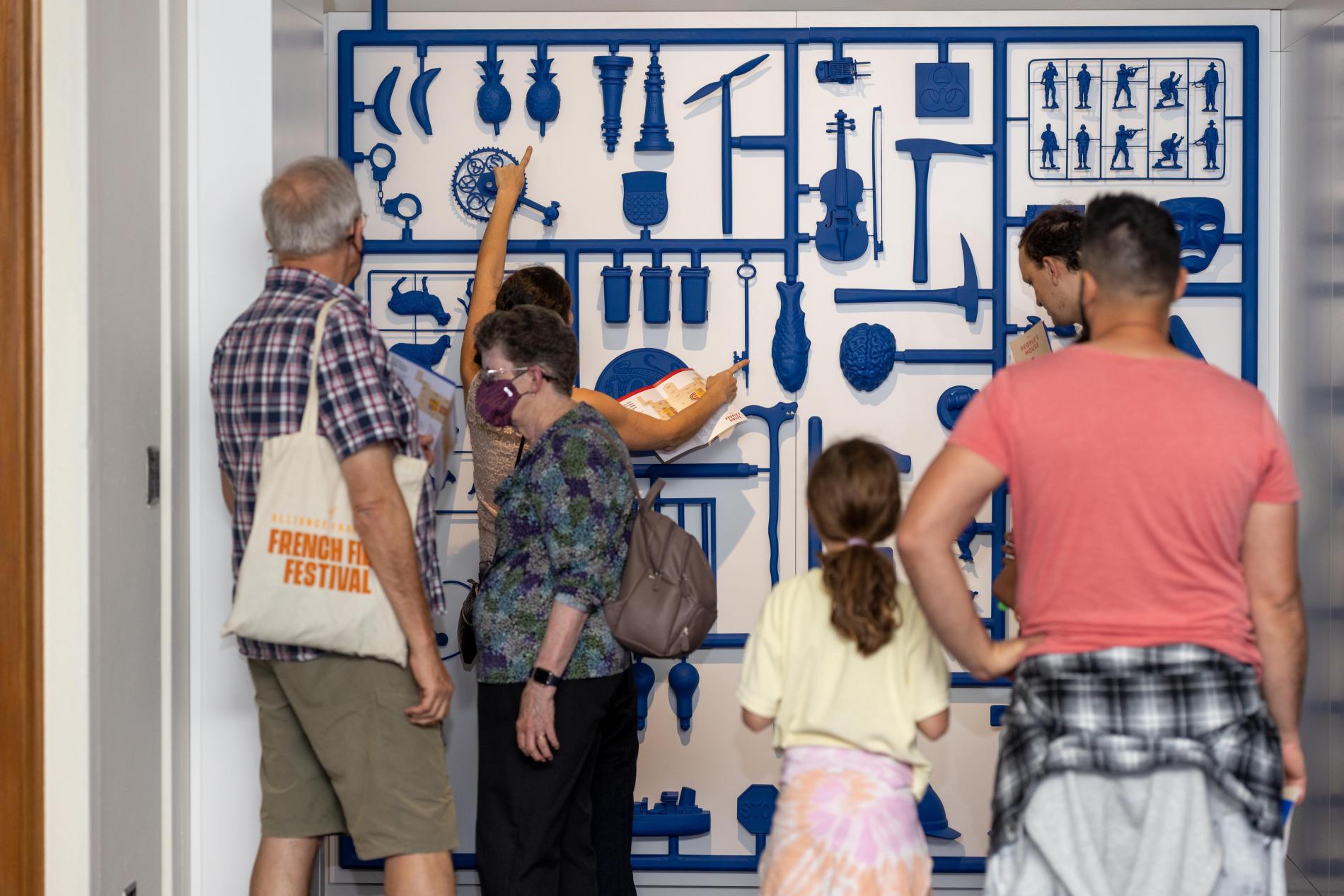 A group of visitors gather in front of a wall with a large blue installation that looks like the plastic pieces of a board game.