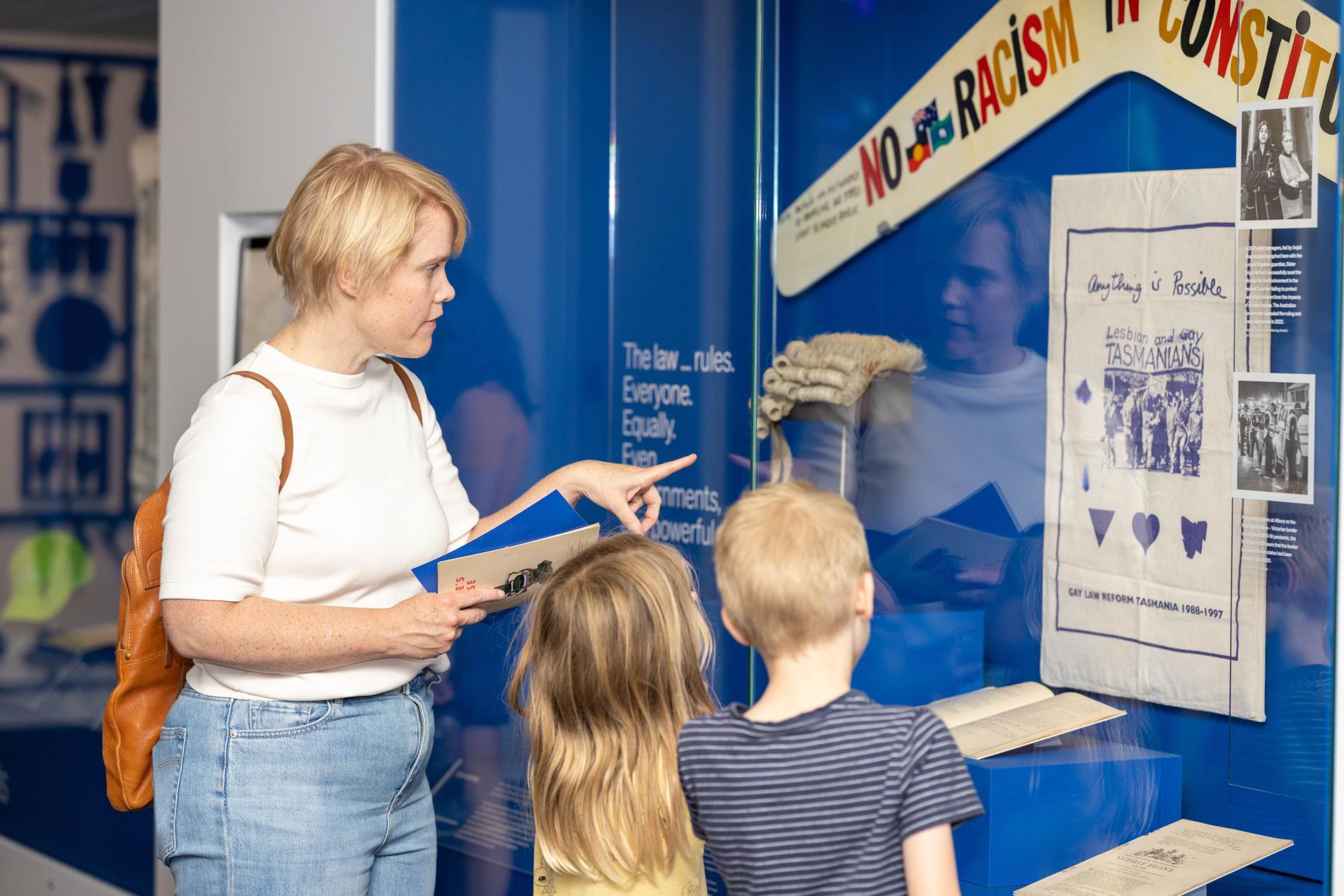 A woman standing with a boy and a girl points to a glass case with a boomerang with the words no racism in the constitution on it.