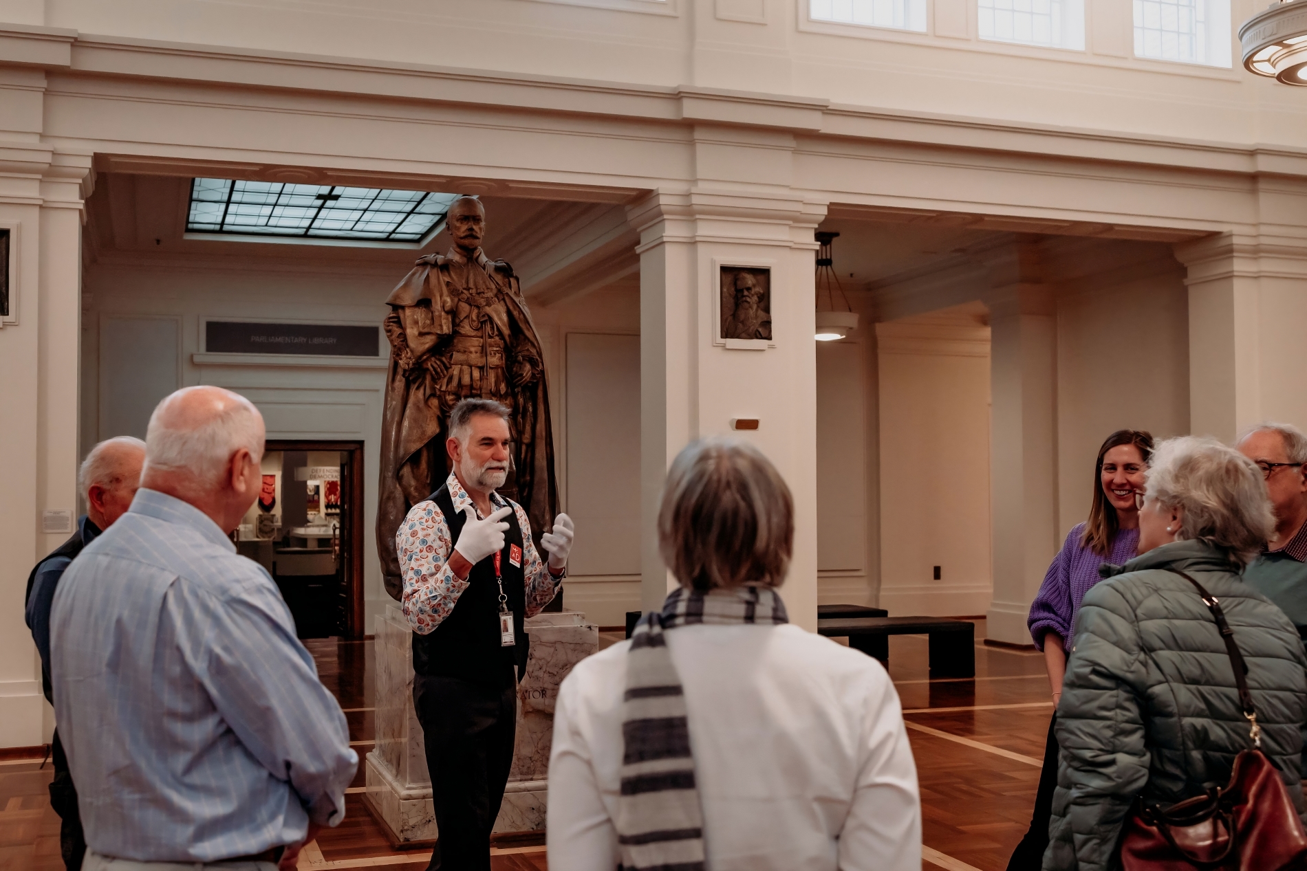 A tour group stand in a circle around a tour guide who wears white gloves and stands in front of a bronze statue.