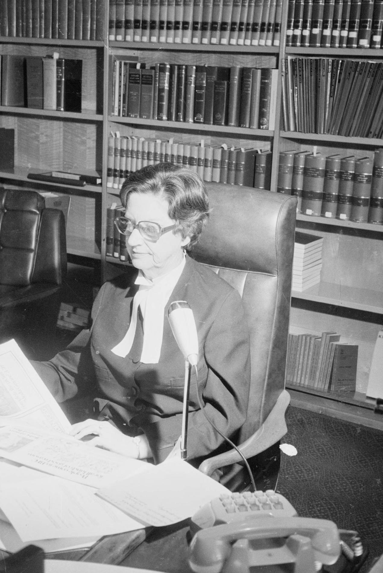 A woman, Justice Roma Mitchell sits at a desk covered in papers, in front of a bookshelf filled with bound volumes.