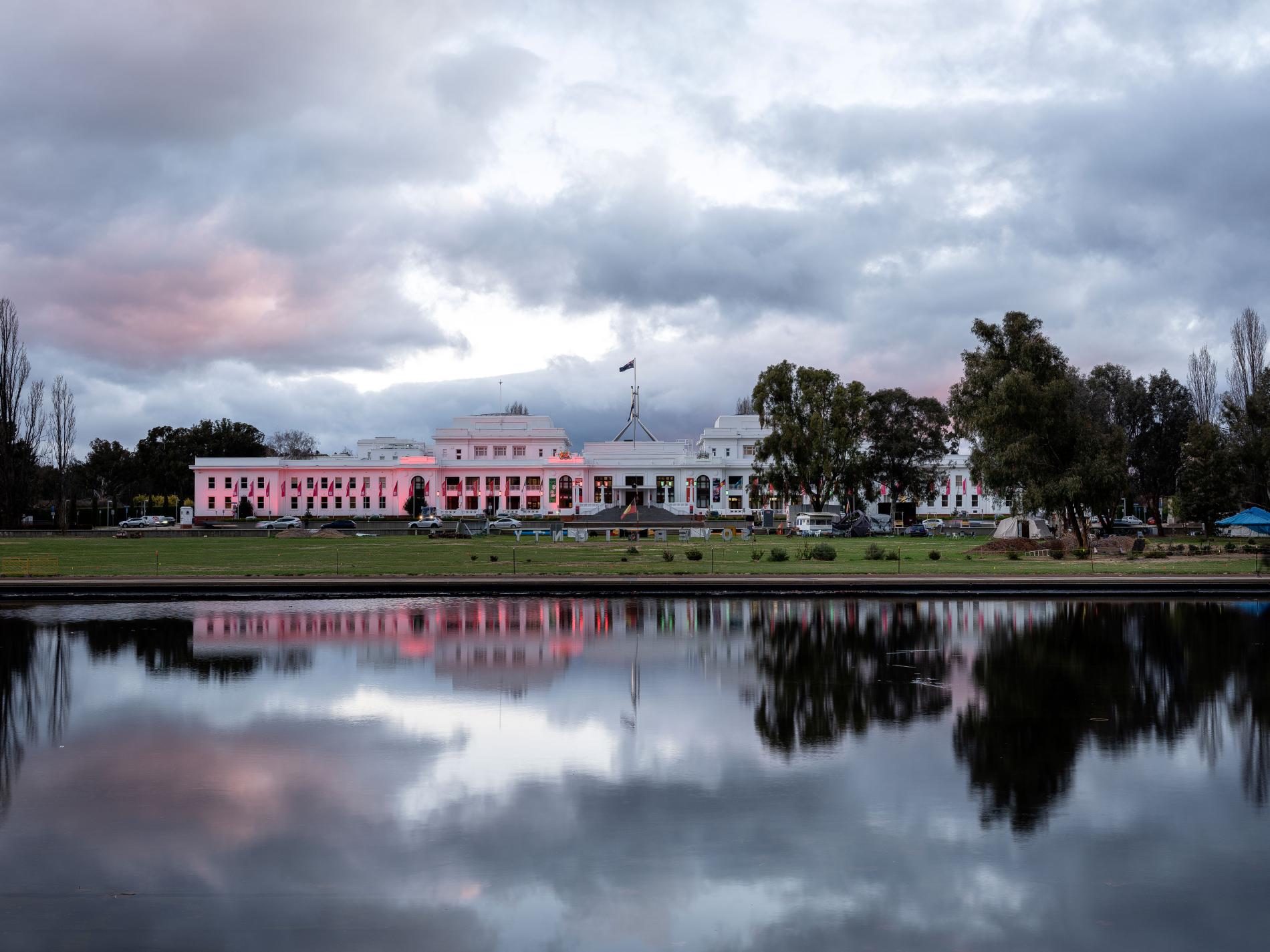 A large white building with the Australian flag on top, with a body of water in front that shows the reflection. The image is at dusk. 