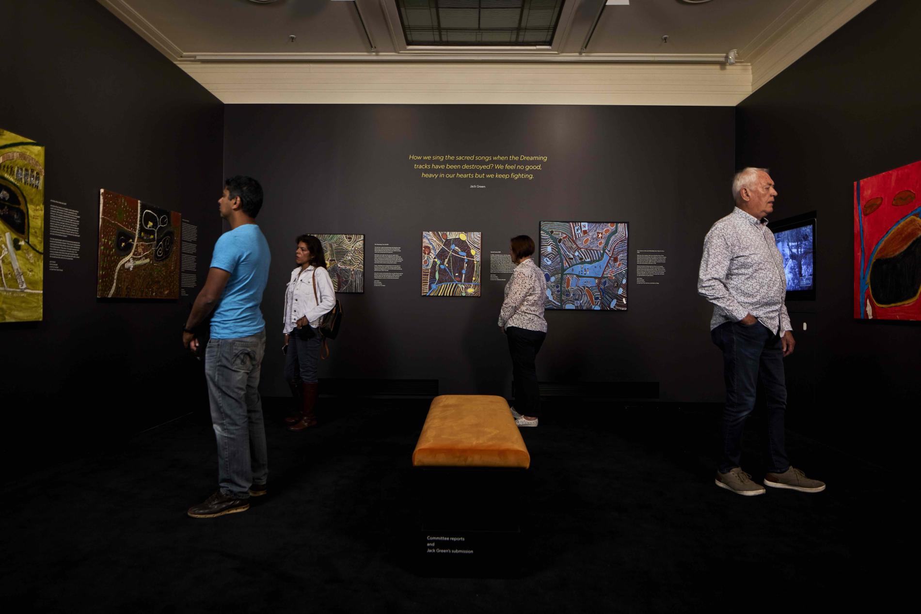 4 people stand in a museum room looking at paintings on the wall by artist Jack Green.