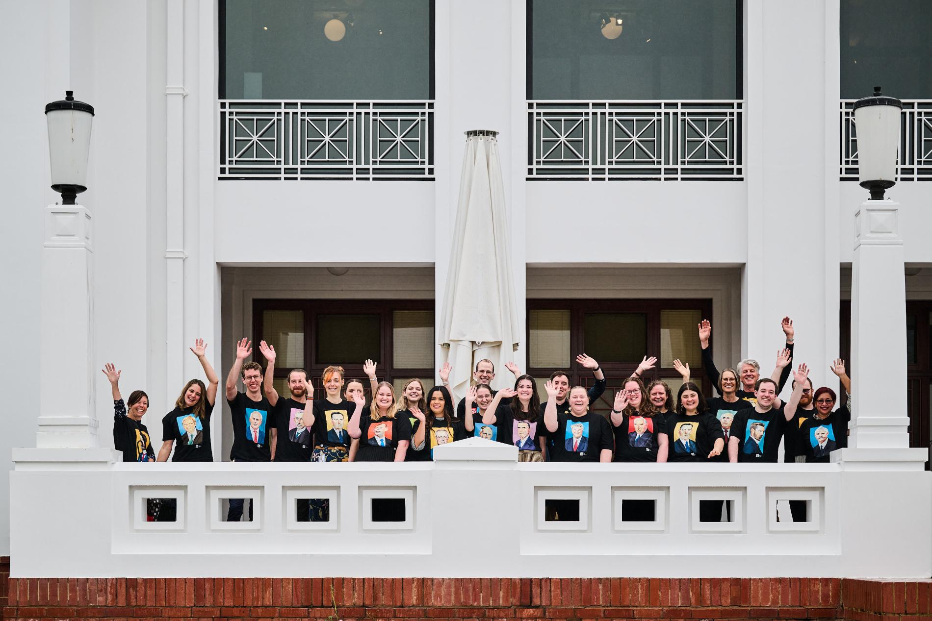 A group of people in black T-shirts with illustrations of prime ministers on them wave from a white terrace balcony.