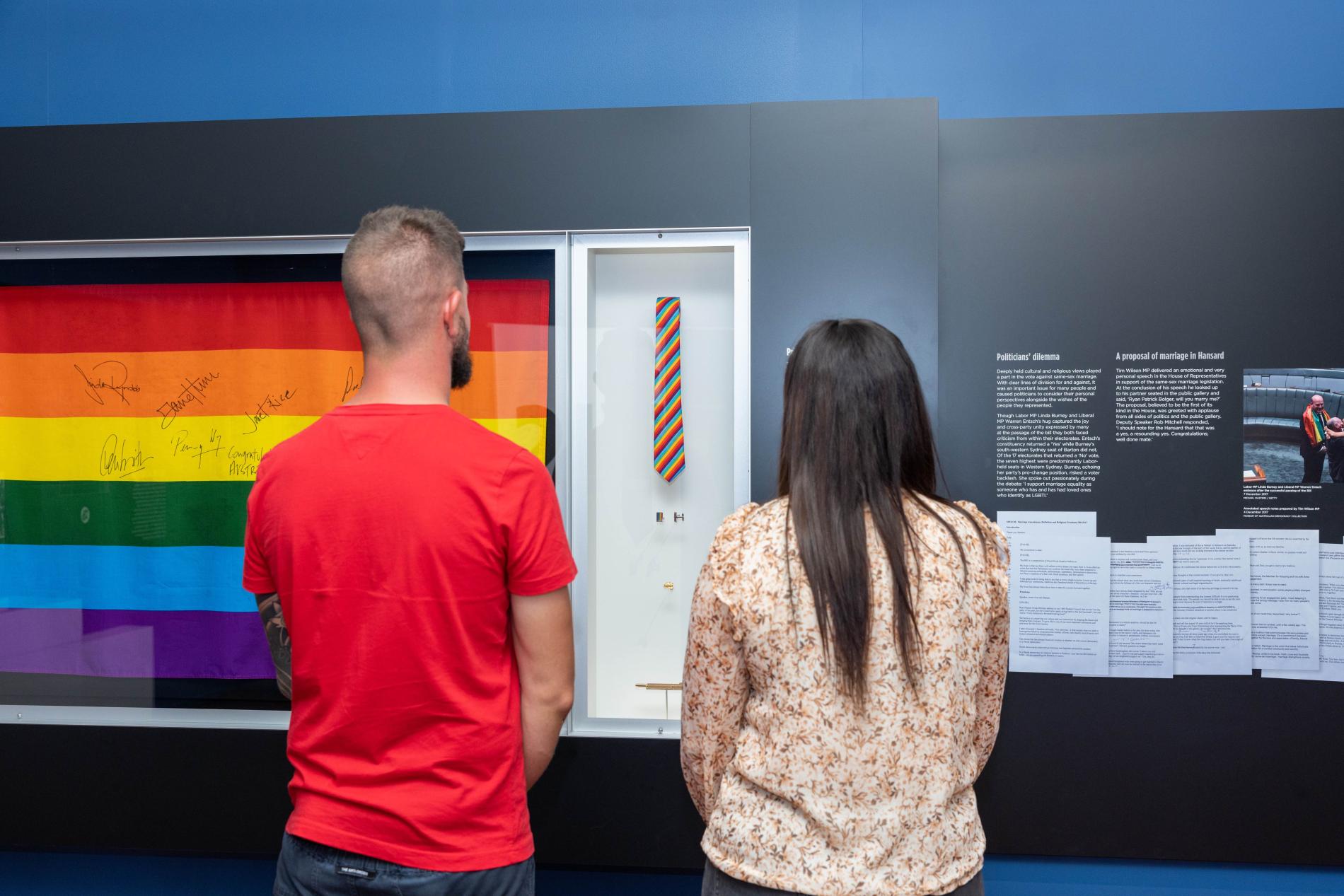 A man and a woman stand in front of a display case with a rainbow flag with signatures on it. 