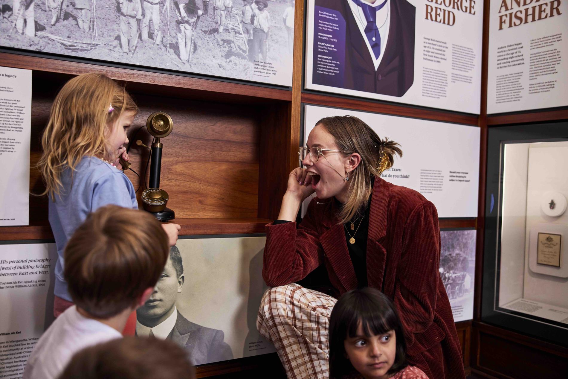 A woman smiles at a child holding a telephone to her ear in a museum exhibition with panels about prime ministers. 