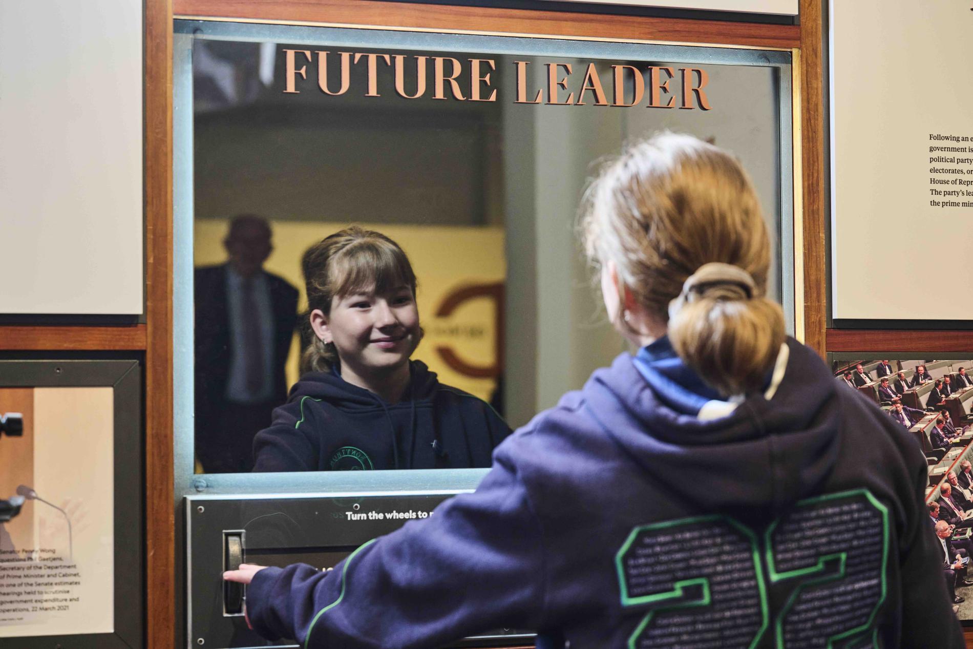 A primary school girl wearing her school jumper looks into a mirror with the words future leader on it. 