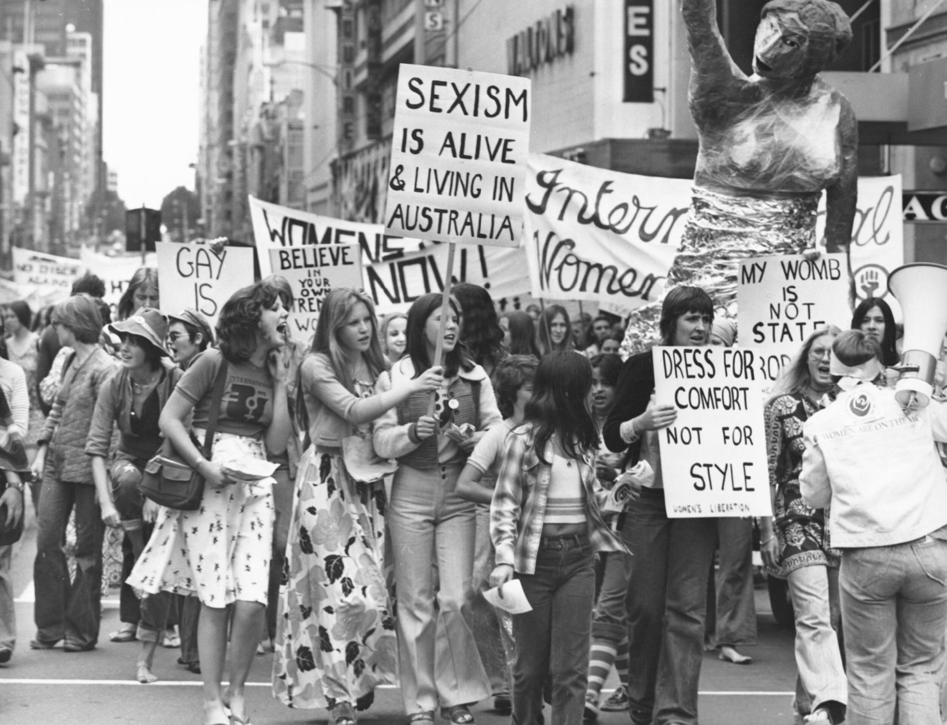 A black and white photo of a group of women holding protest signs in 1975. Signs say 'sexism is alive and well in Australia' and 'dress for comfort not style'.