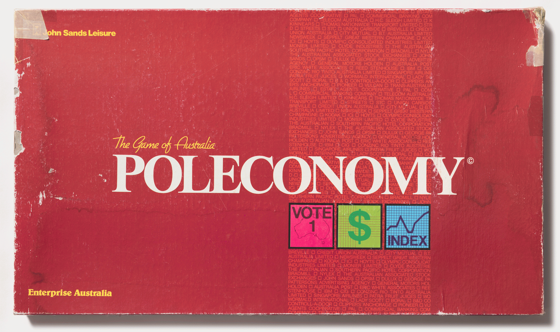 A dark red board game box with the edges slightly scuffed some masking tape around the corners. The word 'Poleconomy' in large white capitals is on the centre of the box. Three square images are in a row beneath it. One is a pink square with the words Vote 1, another is a green square with a dollar sign, a blue square has the word inmd