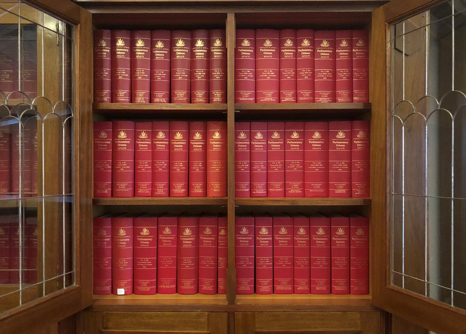 A wooden shelf with opened glass doors holds three rows of red bound volumes labelled 'Parliamentary Debates; Senate' on the spines.