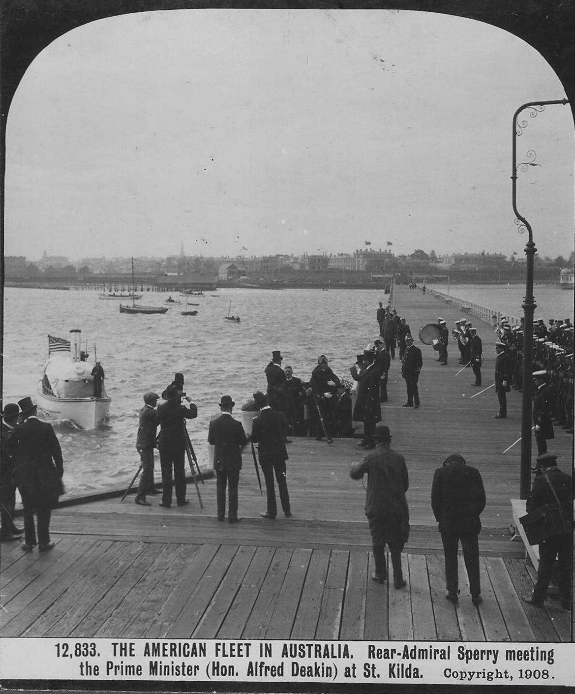 An elevated view from above and behind small clusters of people who stand on a wooden pier. They are looking out at a boat that is headed towards the pier.  The label reads "The American Fleet in Australia. Rear-Admiral Sperry meeting the Prime Minister (Hon. Alfred Deakin) at St. Kilda." 1908.