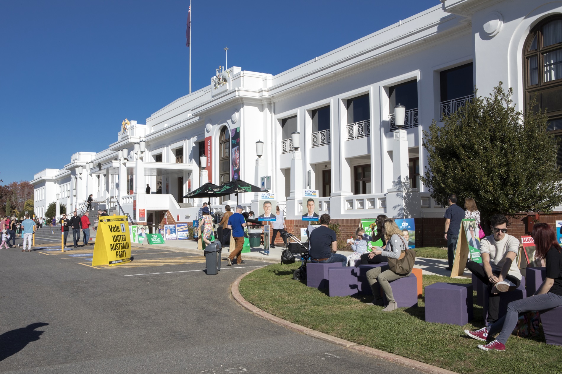 Many colourful signs and placards displaying political candidates, as people enter and leave Old Parliament House, with some sitting on purple blocks on the front lawn.