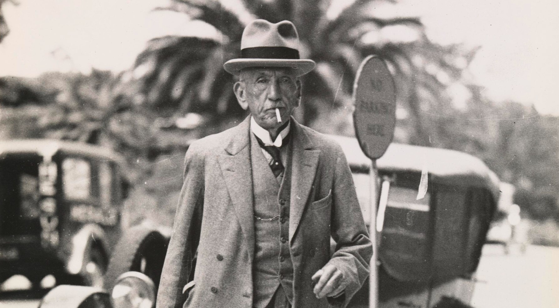 Detail of William Hughes walks down the street wearing a hat and three-piece suit, smoking a cigarette.