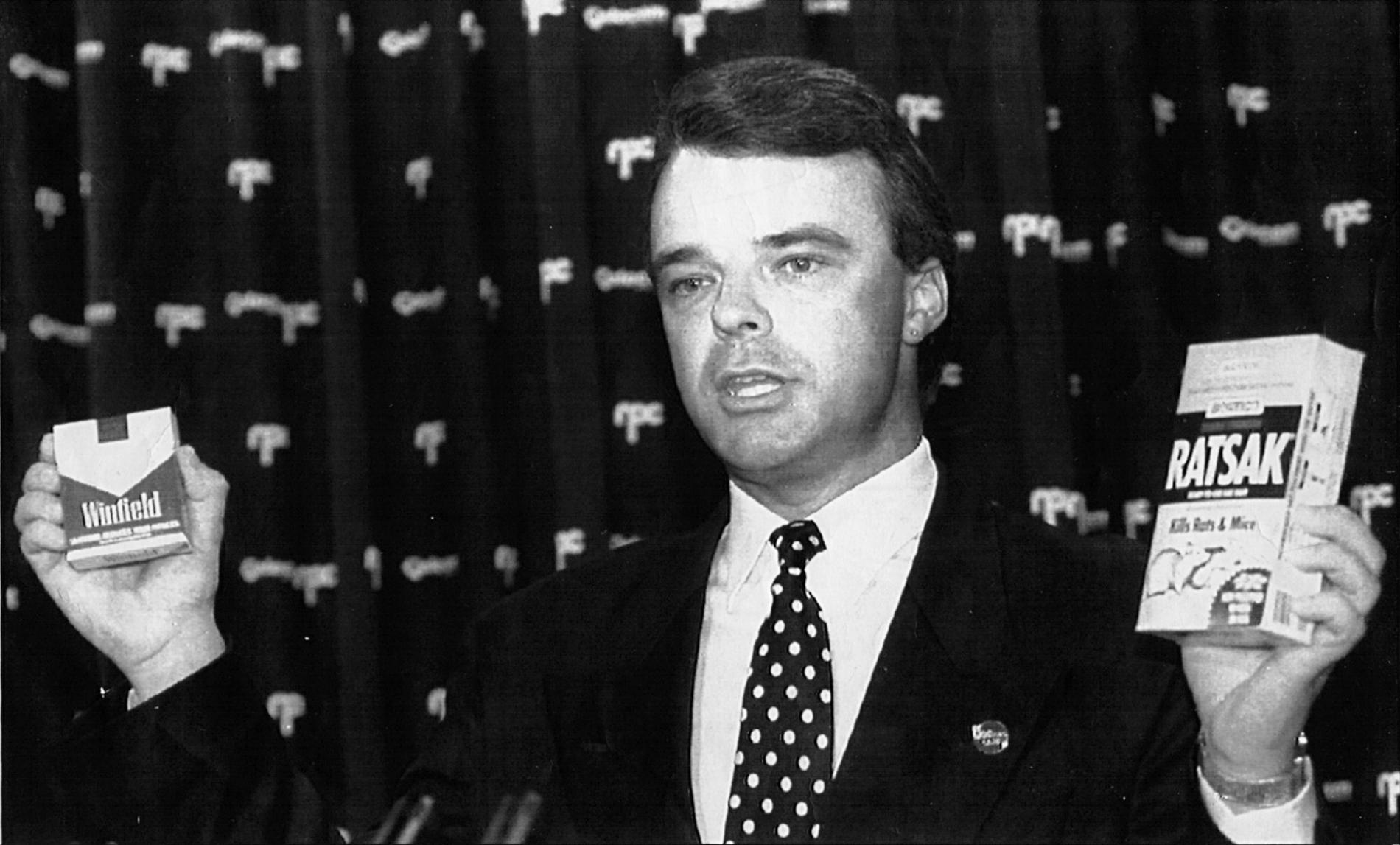 Dr Brendan Nelson, wearing a suit and spotted tie, holding a 'Winfield' cigarette box in one hand and a 'Ratsak' box in the other.