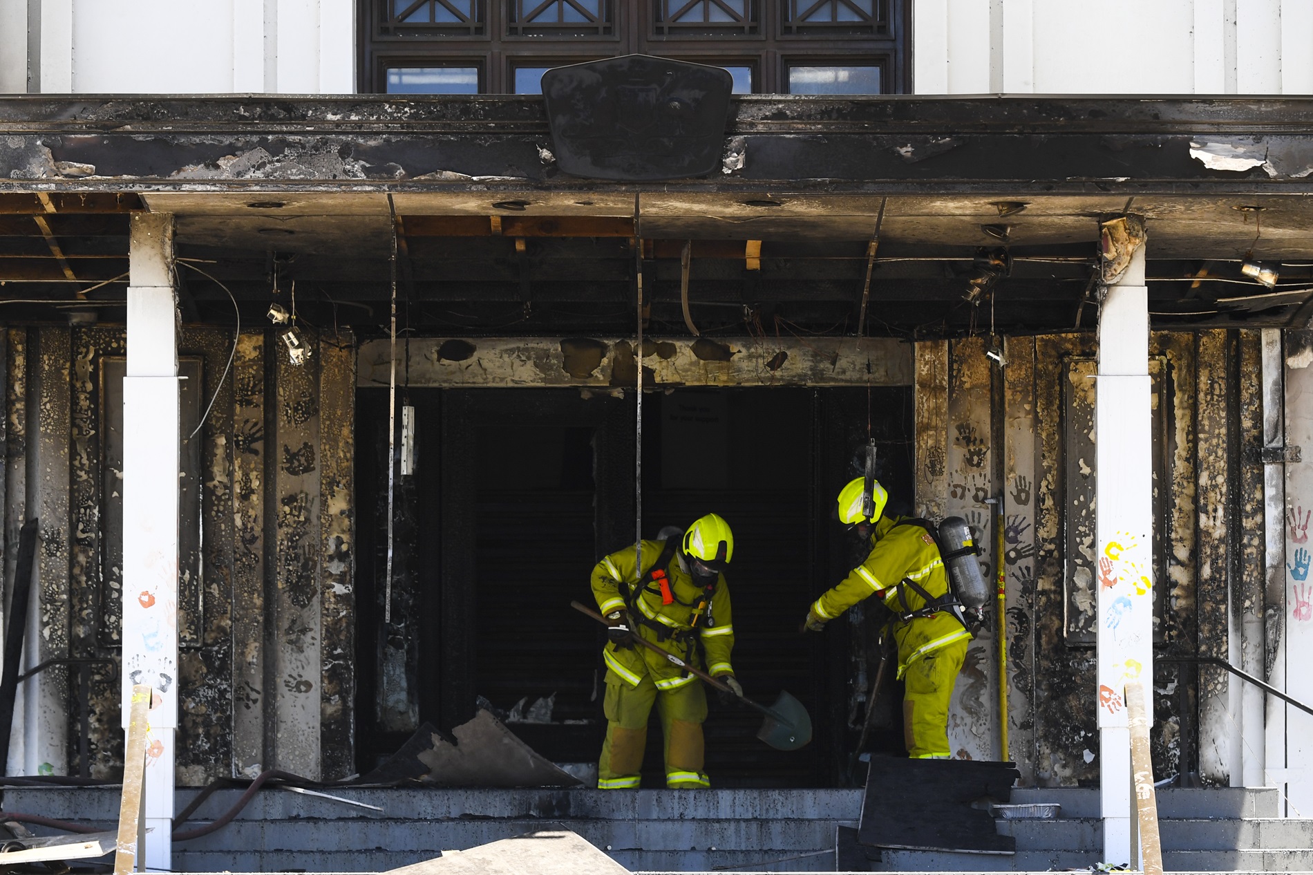 Two firefighters dressed in yellow hi-vis clothing and protective equipment, hold shovels and assess the fire damage to the front entrance of Old Parliament House.