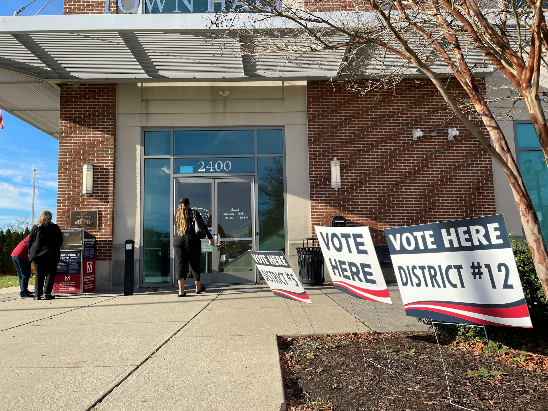 The front of a building with signage that reads 'Vote here', 'District #5' and 'District #12'. One person enters the building, whilst two others gather around a box that reads 'Official election ballot drop box'.