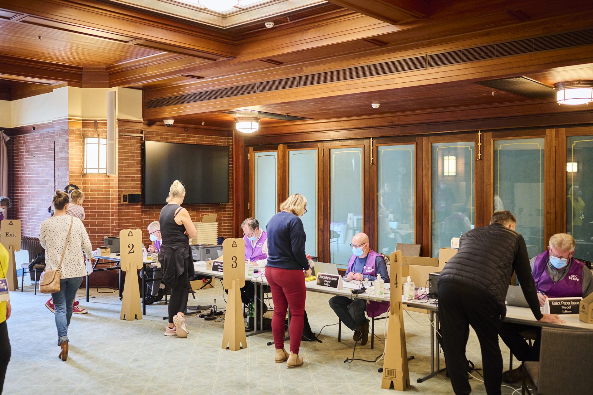 Four Australian Electorial Commission staff sit in a row at a long table wearing face masks and purple vests, and people are lined up, one in front of each staff member, to collect their ballots.