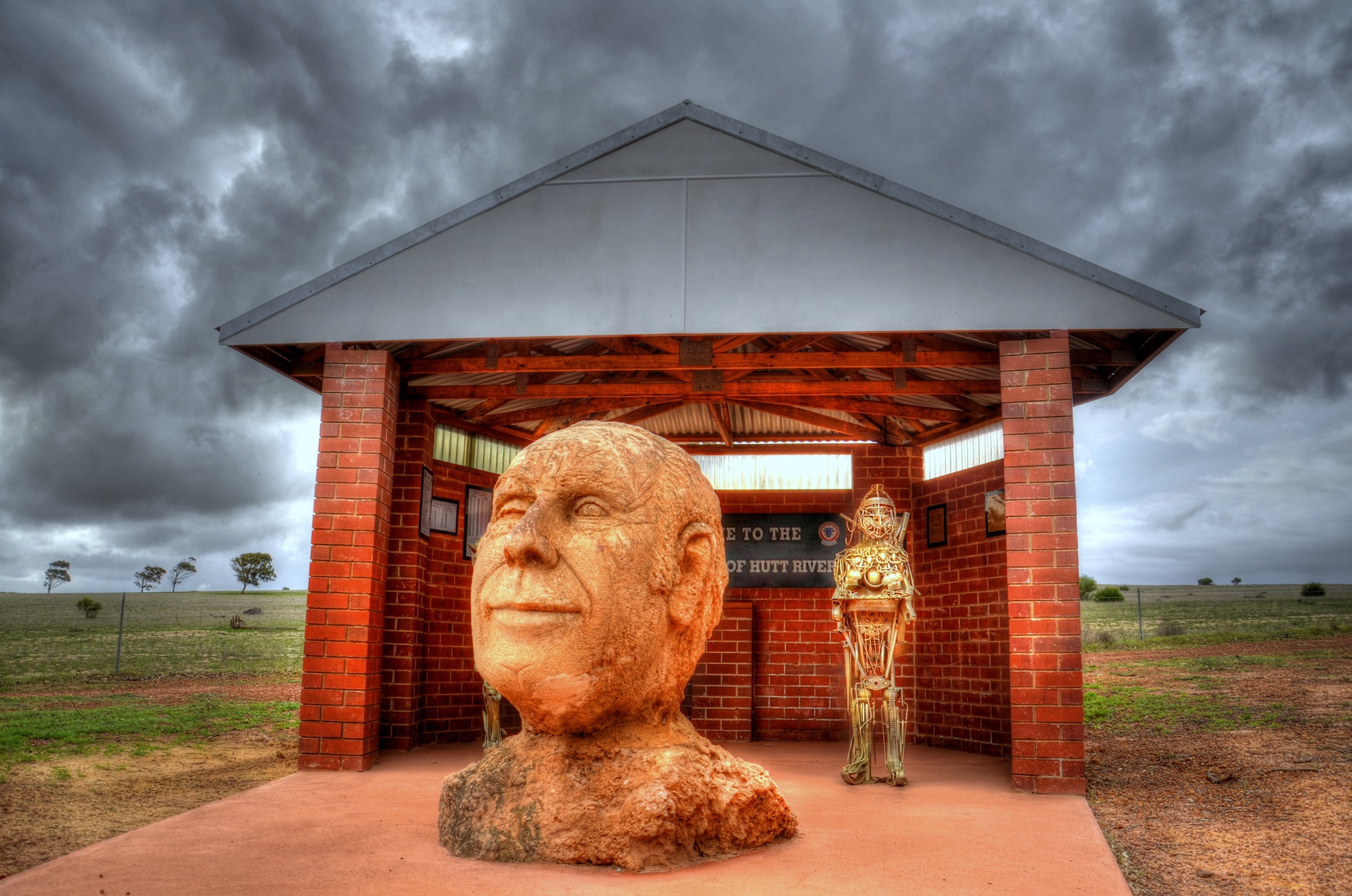 A hand-carved rock bust depicting the head and shoulders of 'Prince' Leonard George Casley, positioned in front of a redbrick shelter in a paddock.