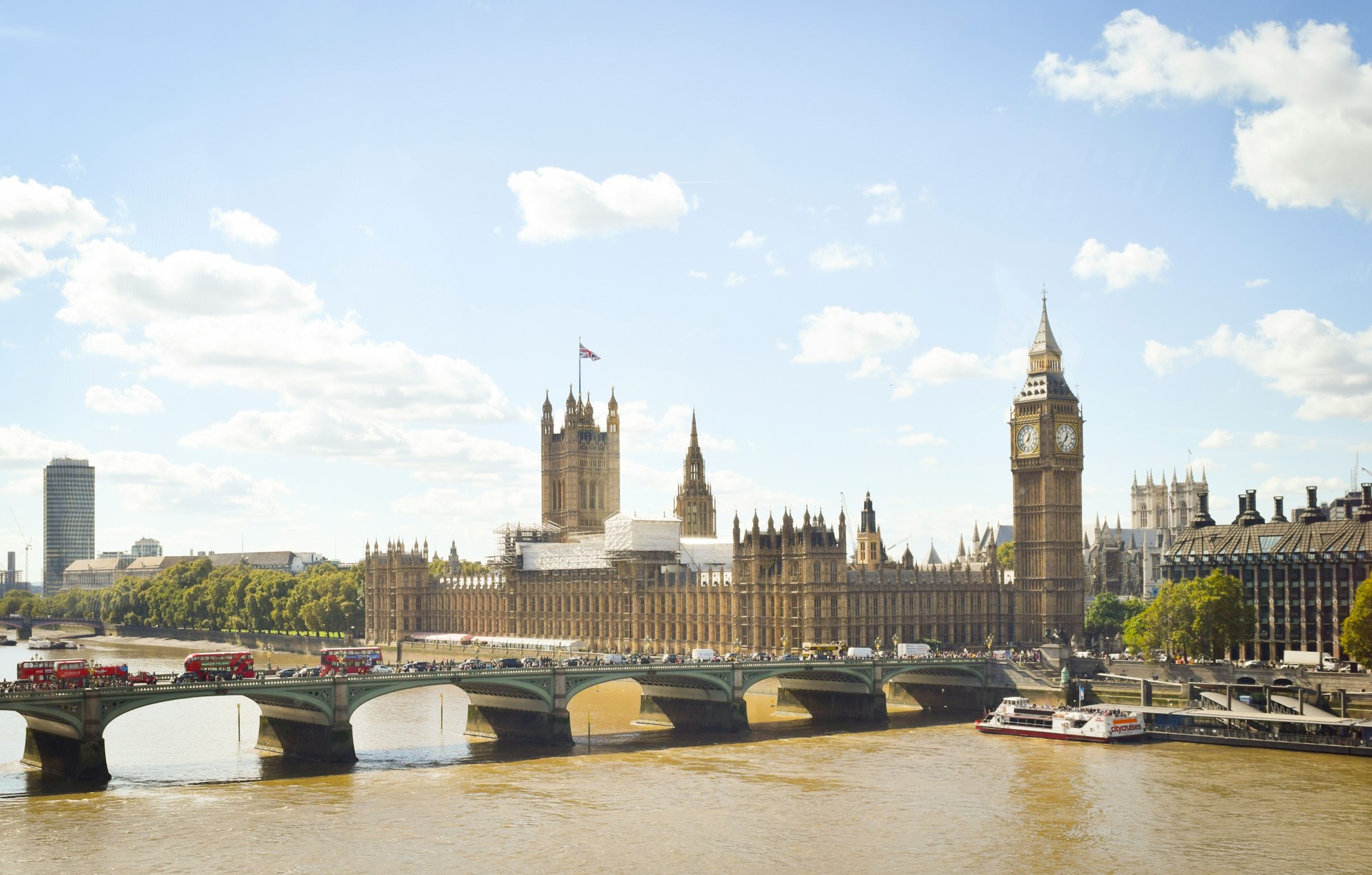 Big Ben and the Palace of Westminster, as viewed from across the river, with red double-decker buses and pedestrians crossing the bridge nearby.