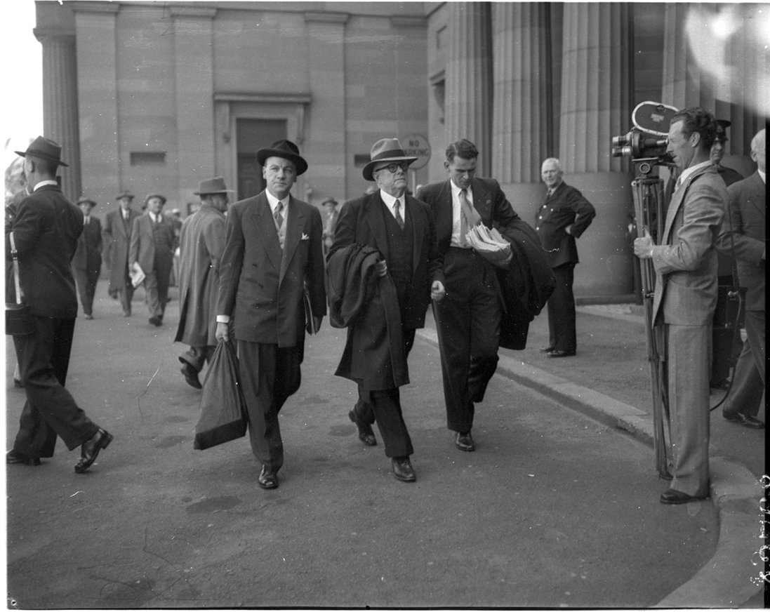 Dr Evatt is flanked by two assistants as he enters the Petrov Royal Commission in Darlinghurst. Men are walking nearby, with a man standing with a camera recording them.