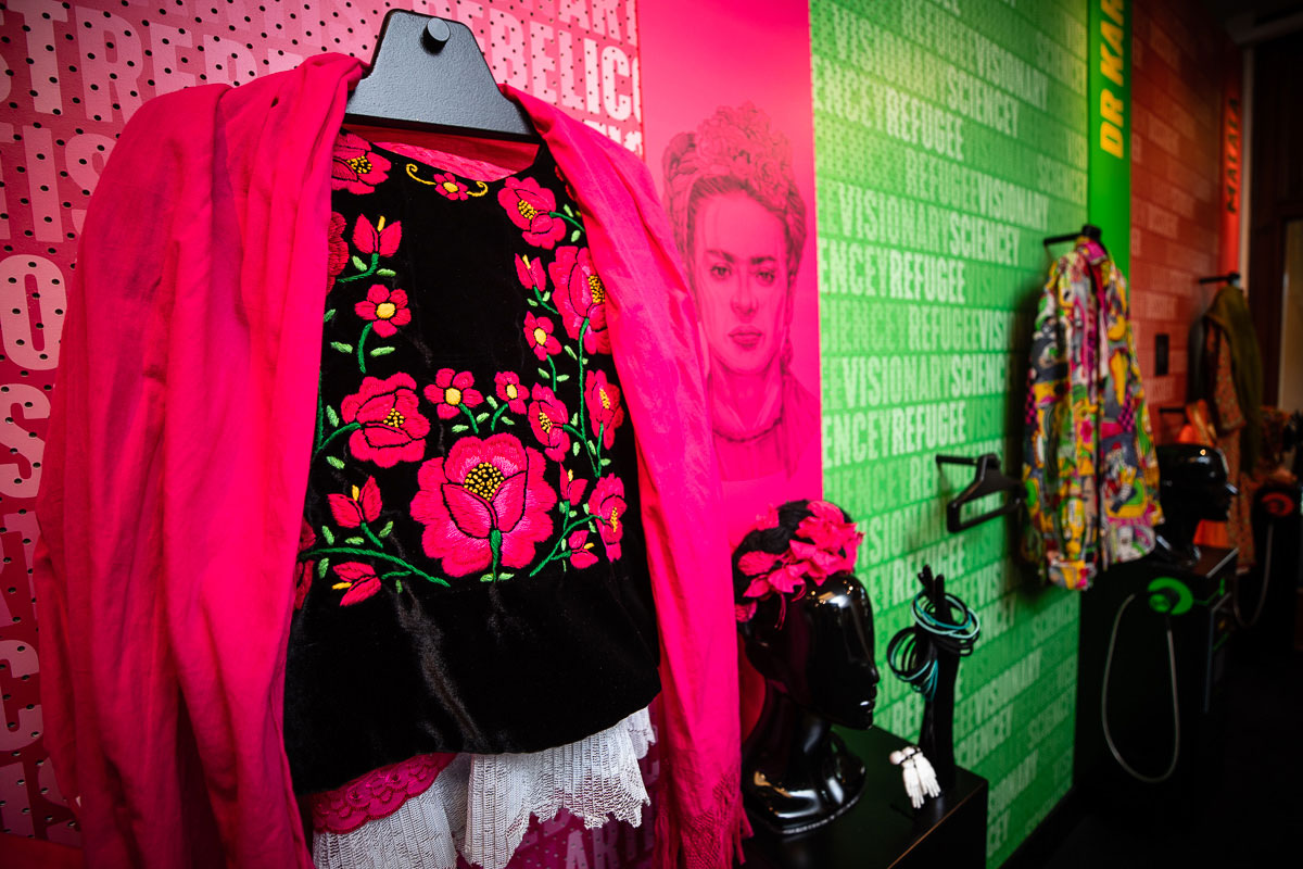 A bright pink coat over a black top with embroidered pink flowers hangs on a pink wall with an image of Frida Kahlo next to it in a room with more costumes to dress up in.