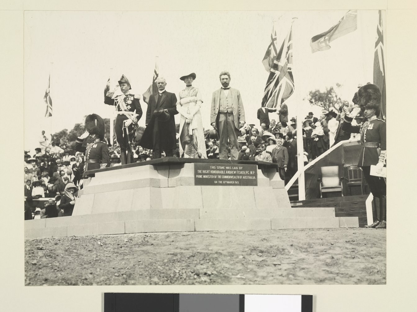 Governor-General Denman, Prime Minister Fisher, Lady Denman and King O'Malley stand on a commemorative stone. British and Australian flags are the in background, with a large crowd behind.