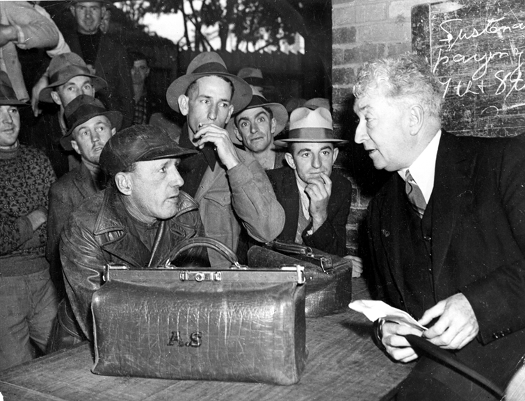 Prime Minister Lyons leans across a desk to chat to wharf workers. A briefcase with the initial A.S. is on the desk. 