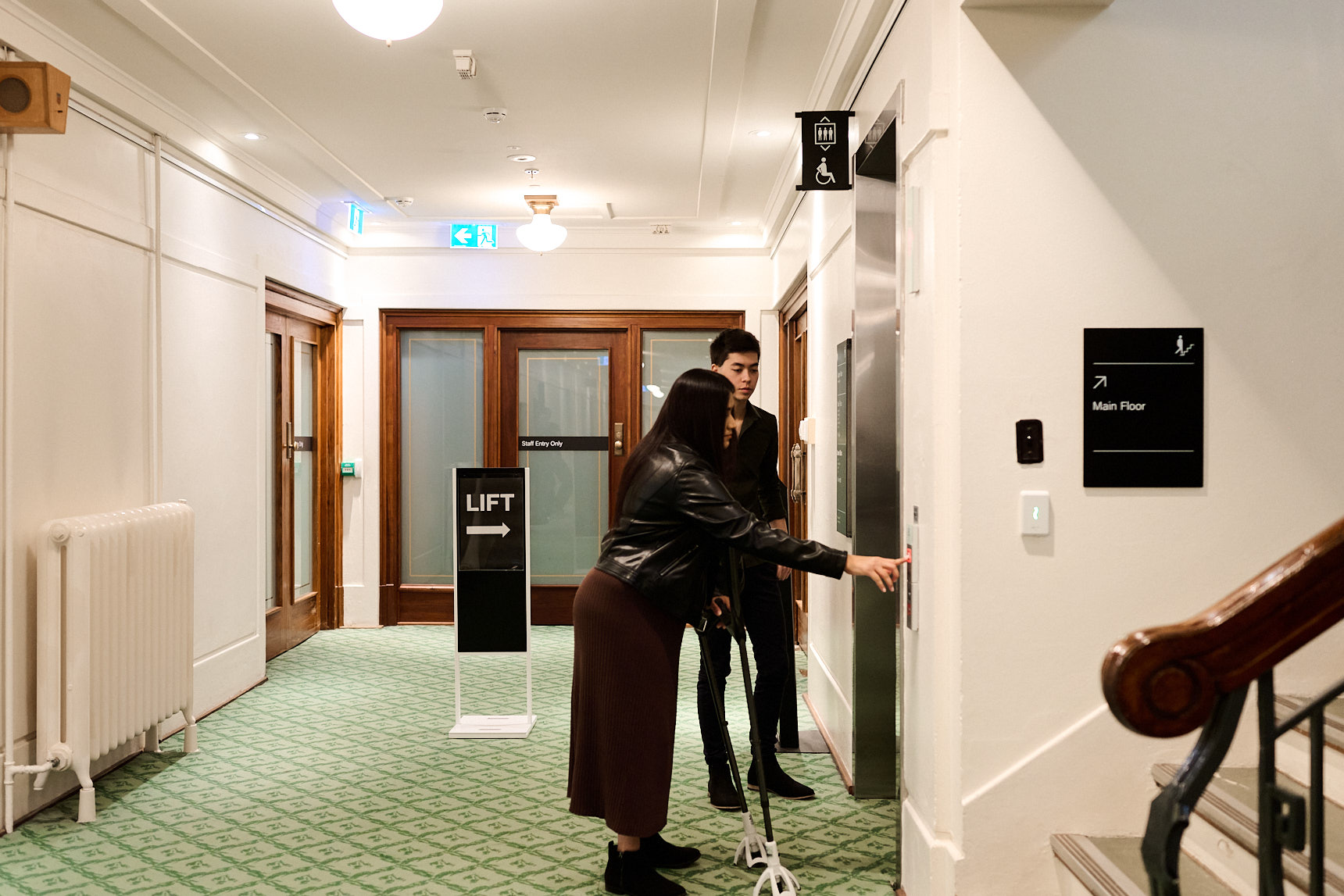 A woman on crutches and a man stand outside a lift on green carpet in Old Parliament House.