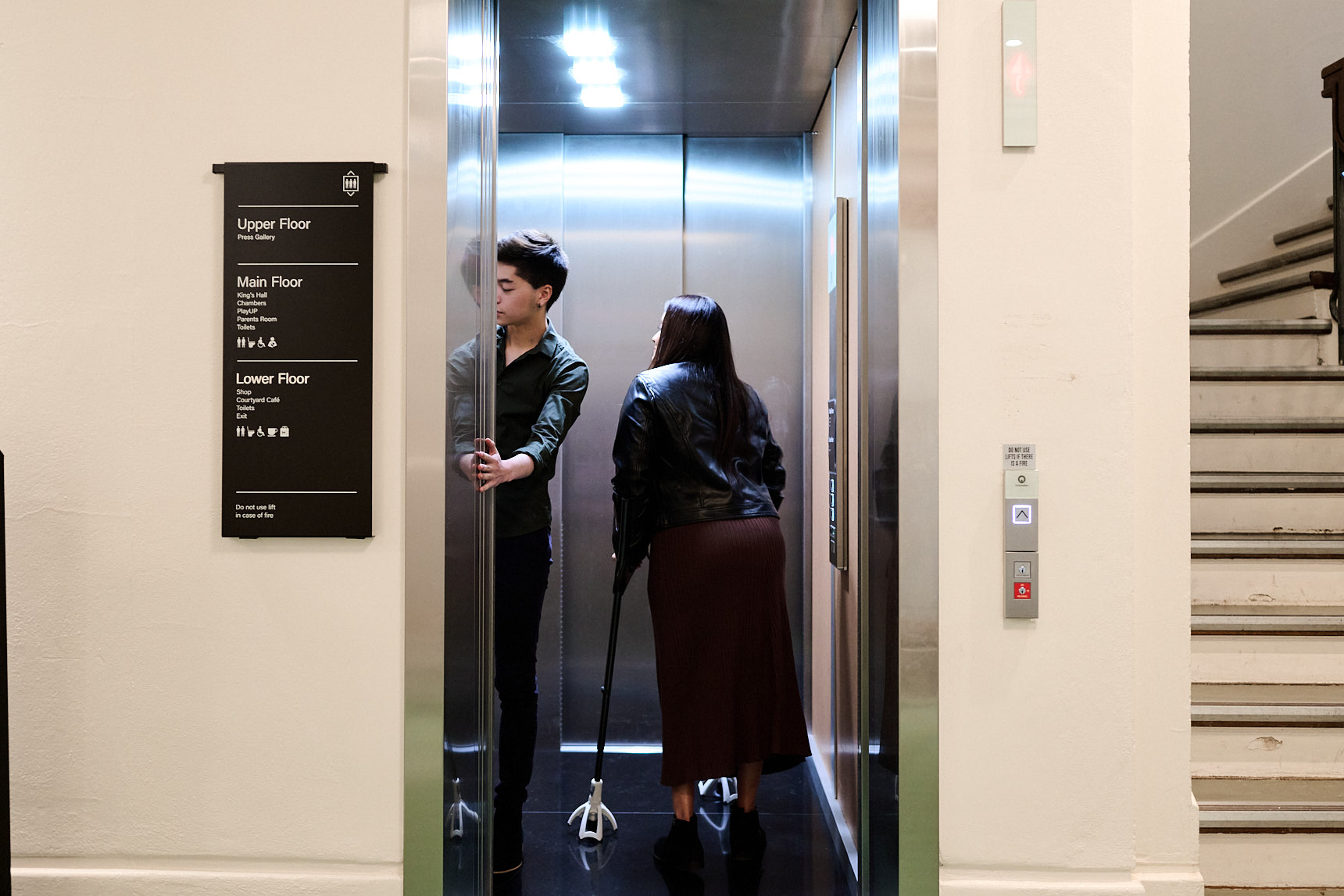 A man holds the door for a woman on crutches inside a lift at Old Parliament House, there is a sign outside the lift for the different floors of the building.