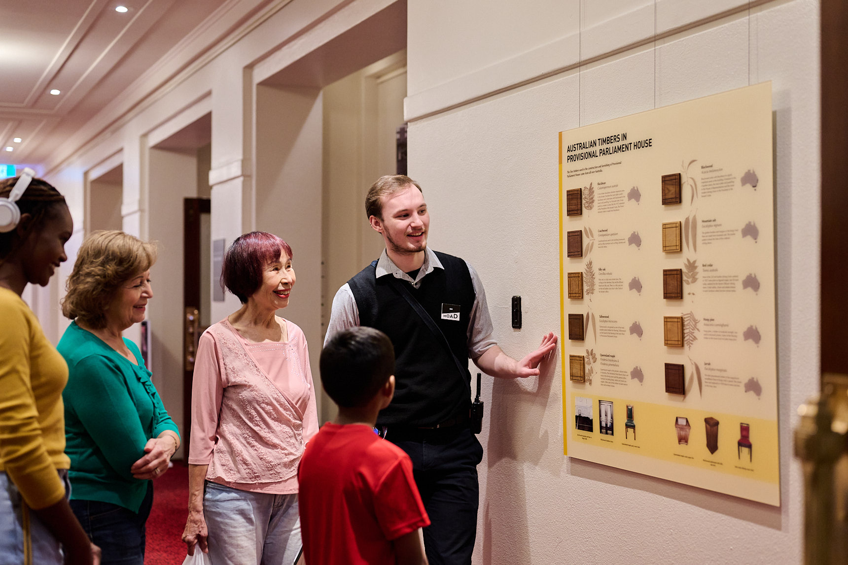 A Museum staff member and a group of people in brightly coloured tops stand in a hallway with red carpet looking at a board displaying Australian timbers used in Old Parliament House.