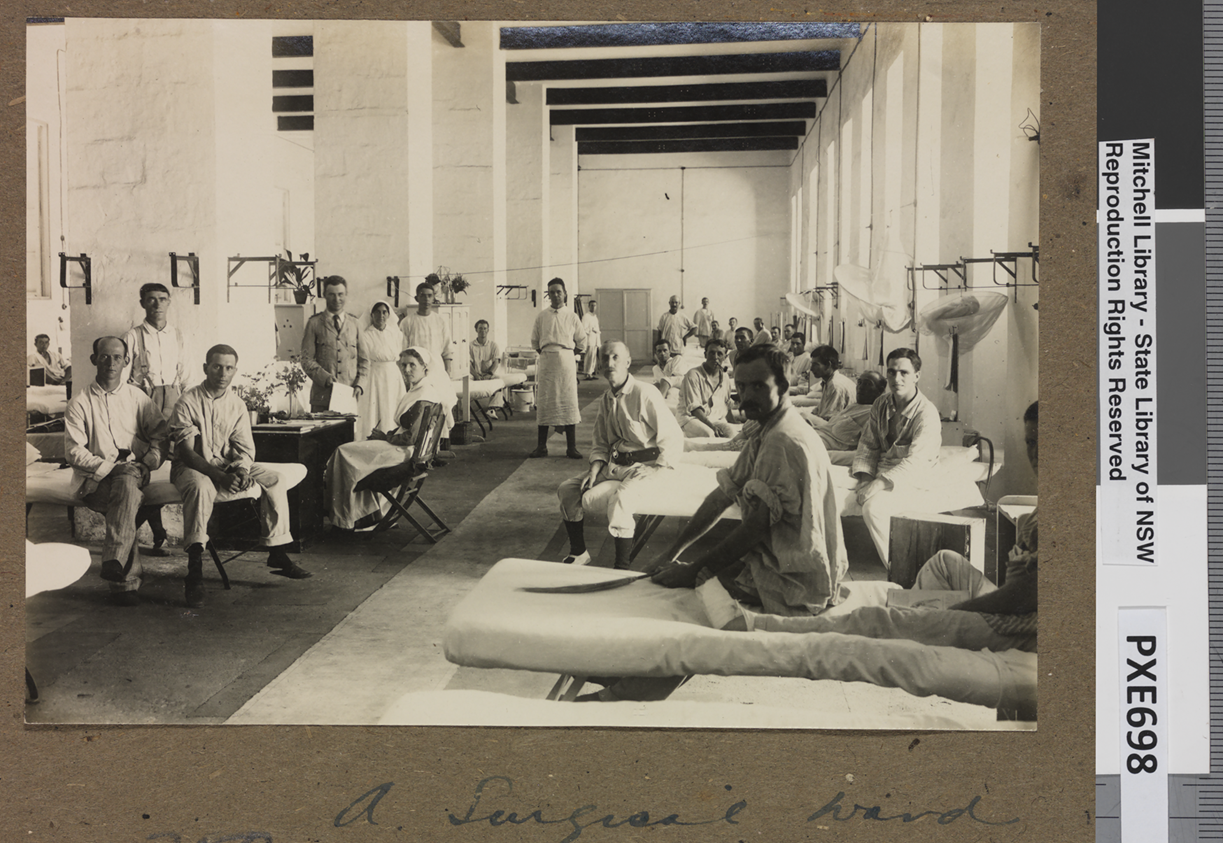 A surgical ward in Egypt during WWII with rows of beds with men looking at the camera. Earle Page is standing holding a piece of paper, next to a nurse. 